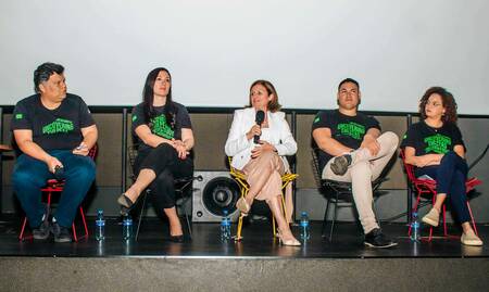 Five individuals sit in chairs on a stage in front of a large screen. A woman in a white blazer speaks into a microphone.  All individuals wear black t-shirts with neon green "Uncovering the Past" text, except the speaker, who is in business attire.