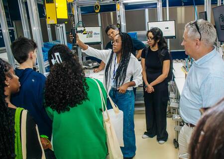 A woman gestures toward a robotic arm in a lab setting.  A diverse group of students and a man in a light blue checked shirt observe.  Computer monitors and equipment are visible in the background.