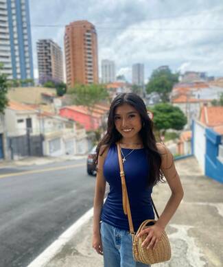 Smiling young woman in blue top & jeans, gold cross necklace, holding a woven bag, stands on a hilly urban street. Background shows diverse buildings with colorful walls & tiled roofs, and taller city structures under a cloudy sky.