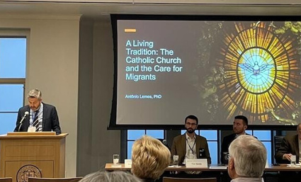 Man in suit speaks at a Notre Dame podium. Screen displays 'The Catholic Church and the Care for Migrants' with a dove.