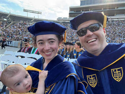 Two adults in blue doctoral robes, one holding a baby, smile at graduation in Notre Dame Stadium.