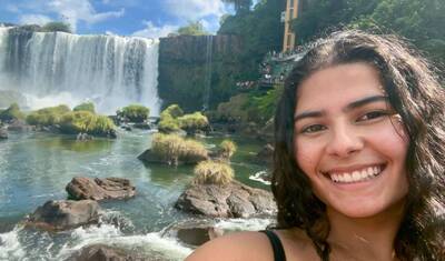 Young woman smiles in a selfie with the vast Iguazu Falls, river, green islands, and an observation deck behind her.
